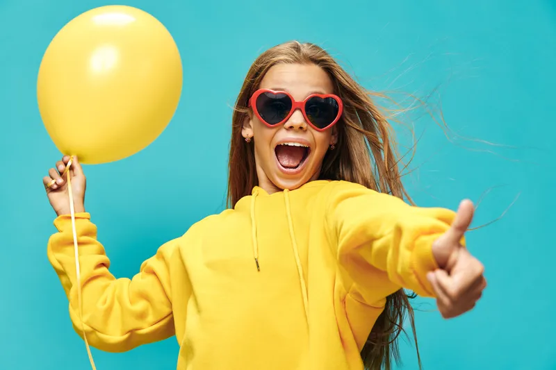 Young woman in yellow hoodie holding yellow balloon with excited expression and thumb up against blue background for celebration and joy.