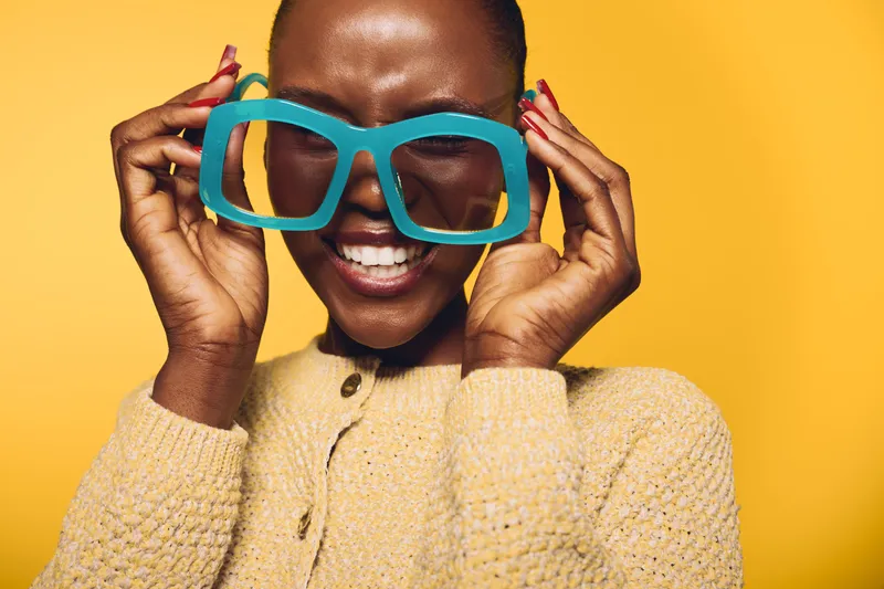 Happy African woman with dark skin wearing oversized blue glasses and beige sweater smiles with closed eyes on yellow background. Concept of joy, fashion, and positive mood.