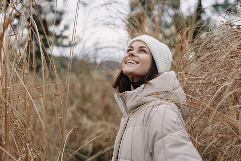 Woman in a warm coat and beanie outdoors, smiling among tall grasses in a chilly field, enjoying nature and fresh air and a peaceful landscape.