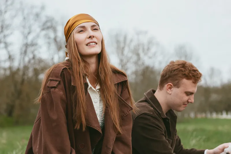 An outdoors lifestyle portrait showing a couple in a windy countryside setting, a woman in a brown coat and head scarf, with a man nearby, capturing candid, relaxed fashion.