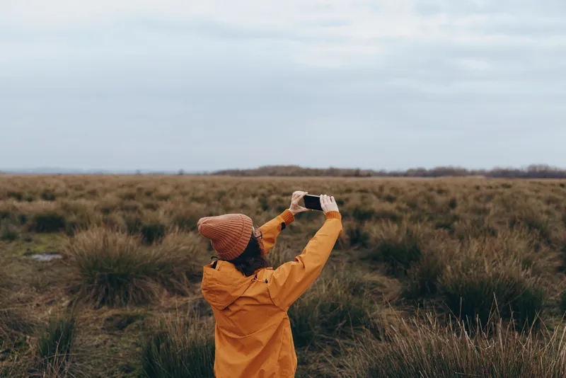 Woman in yellow raincoat and straw hat stands in grassy field outdoors, lifting hands to catch wind, lifestyle portrait, nature landscape, calm mood, soft daylight