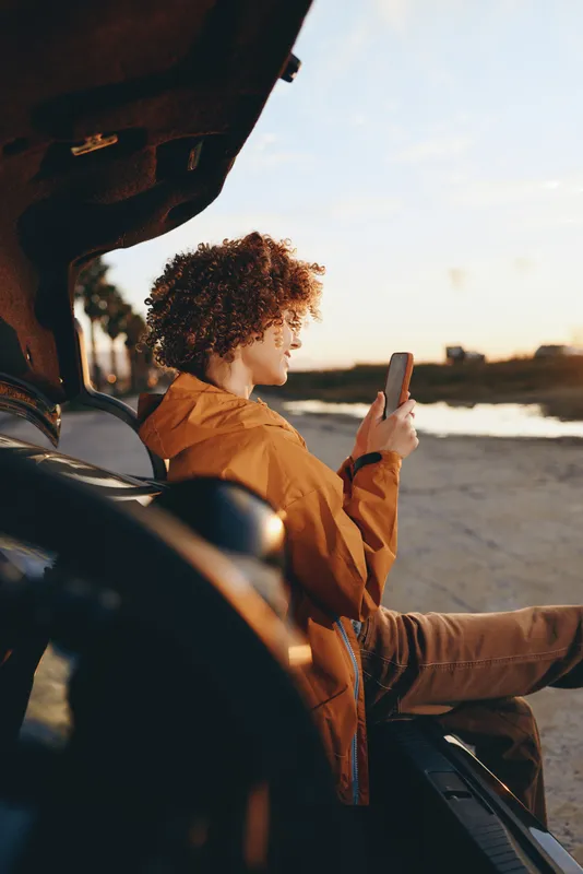 Woman lifestyle in car, passenger seat, curly hair, using smartphone while traveling at sunset, roadside view, warm light, casual travel concept, modern mobility on road trip, open window