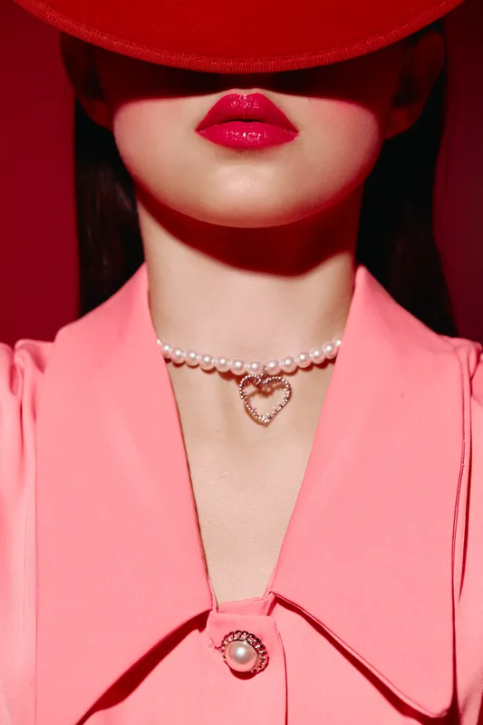 Female model in pink outfit wears bold red lipstick and pearl necklace with pendant heart. Red hat shading eyes adds mystery. Beauty fashion creative portrait concept.