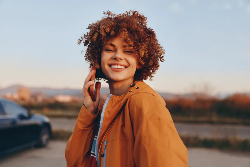 Happy woman with curly hair in an orange jacket holds a smartphone to her ear, smiling outdoors in golden hour sunset light, city riverside background, lifestyle concept for friendly communication
