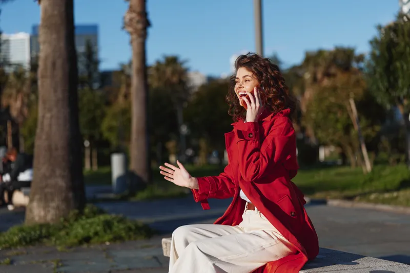 A young woman on the phone sits on a bench in a sunny park, wearing a red coat and cream casual outfit, capturing a candid moment of everyday communication, with curly hair framing a focused