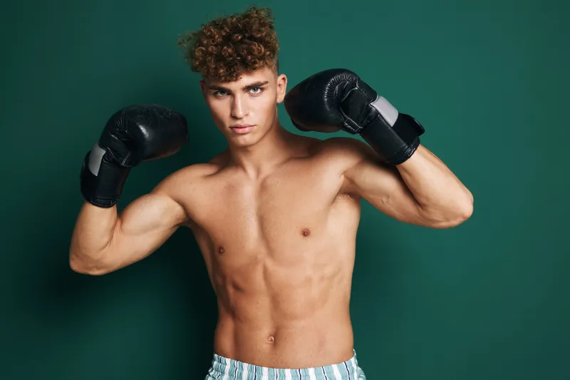 Young muscular man with curly hair wearing boxing gloves posing shirtless against green background. Athlete showing strength and fitness with confident expression in studio lighting.