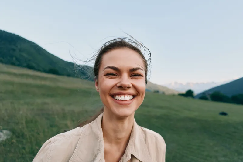 Smiling woman in a beige shirt stands in a green field with distant mountains under a sunny sky, conveying casual outdoor bliss and natural confidence for lifestyle photography ideas and editorial