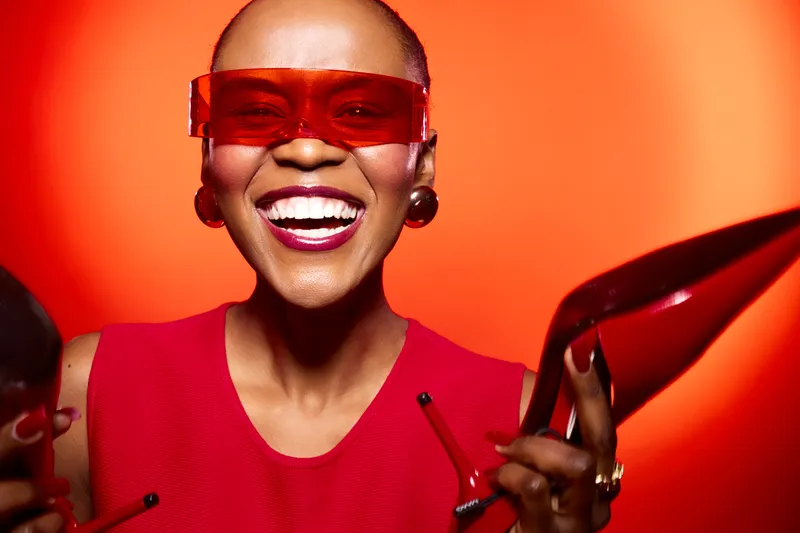 Smiling black woman with dark skin wearing red sleeveless top and red sunglasses holds red high heels on orange background. Concept of fashion, joy, style, and vibrant mood.