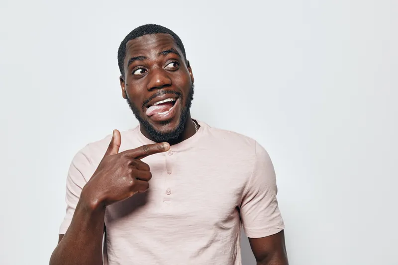 African American man with dark skin pointing finger at himself with surprised expression wearing light pink t shirt on white background. Casual portrait of young black male.