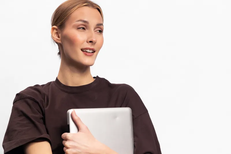 Young woman with laptop in hands looks away and smiles gently, wearing dark casual shirt. Studio portrait on white background. Concept for technology, work, and relaxed mood.