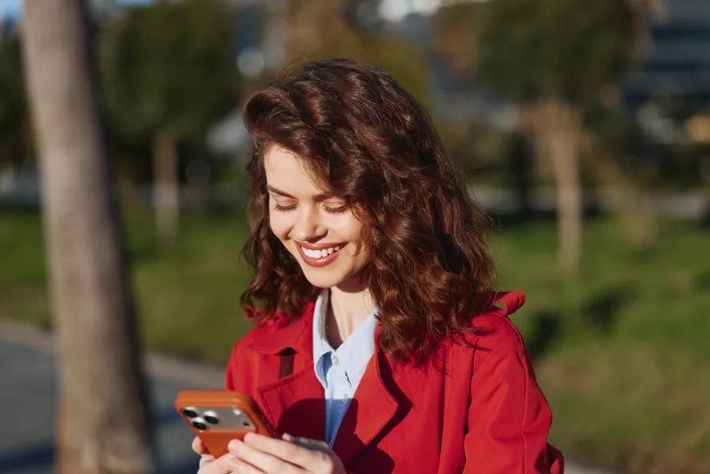 Curly haired woman in a red coat smiles at her smartphone in a sunny outdoor park, capturing a casual moment of everyday life with relaxed pose and soft warm light.