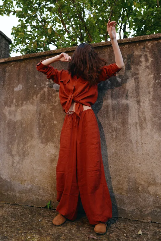 A young woman in a loose red jumpsuit stands with her back to a weathered wall, arms raised, creating a bold urban portrait full of movement and curiosity