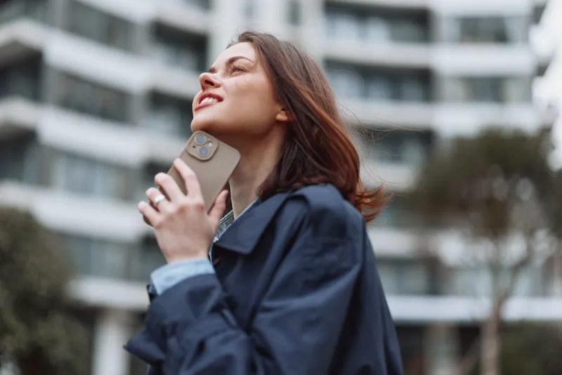 Young woman in a navy jacket chats on a phone outdoors near a modern building, conveying focus and urban lifestyle in a candid moment, with her hair slightly blown and a look upward suggesting