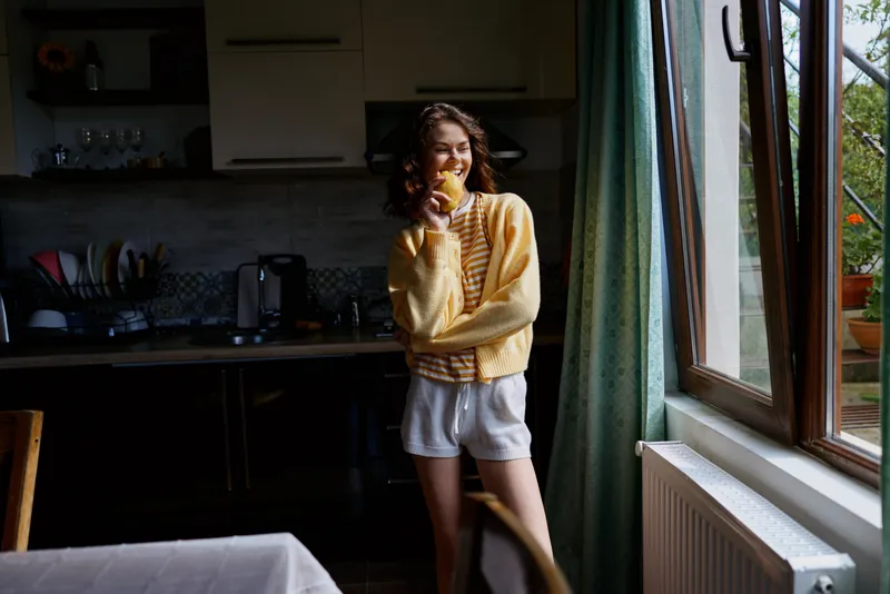 Smiling woman in a yellow sweater stands by a window in a cozy kitchen, enjoying a snack. A casual home lifestyle scene with warm natural light and relaxed mood.