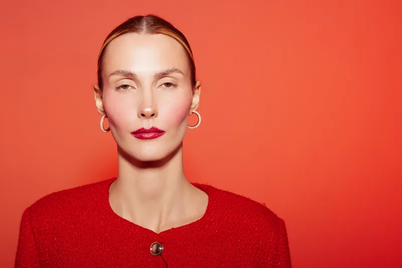 Portrait of confident woman with slicked back hair wearing red jacket and bold lipstick on orange background. Concept of fashion, beauty, and elegance in studio lighting.