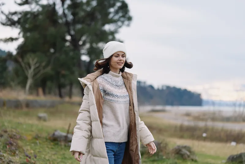 Woman outdoors in a winter coat walking in a park, wearing a sweater and beanie, enjoying a chilly day by the lake with calm landscape and serene nature