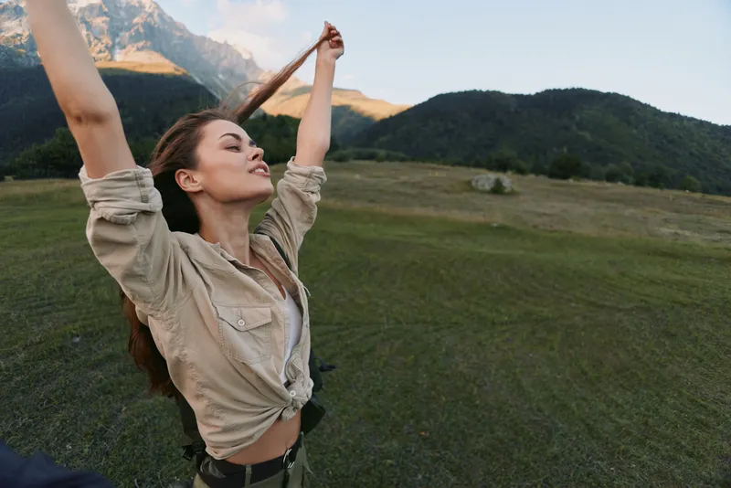 Happy woman outdoors stretching arms up enjoying fresh air in mountain landscape casual clothing natural light peaceful moment
