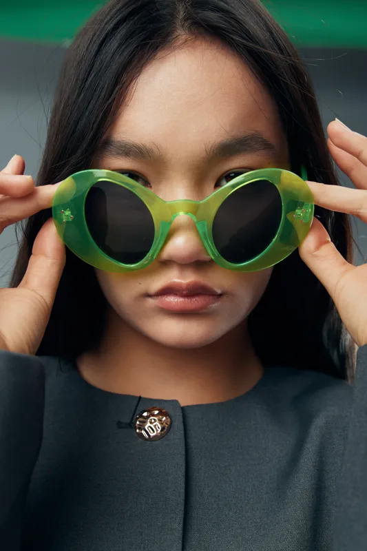 Woman wearing large green sunglasses and dark coat adjusting eyewear with hands, closeup portrait with serious expression and long dark hair outdoors in natural lighting.