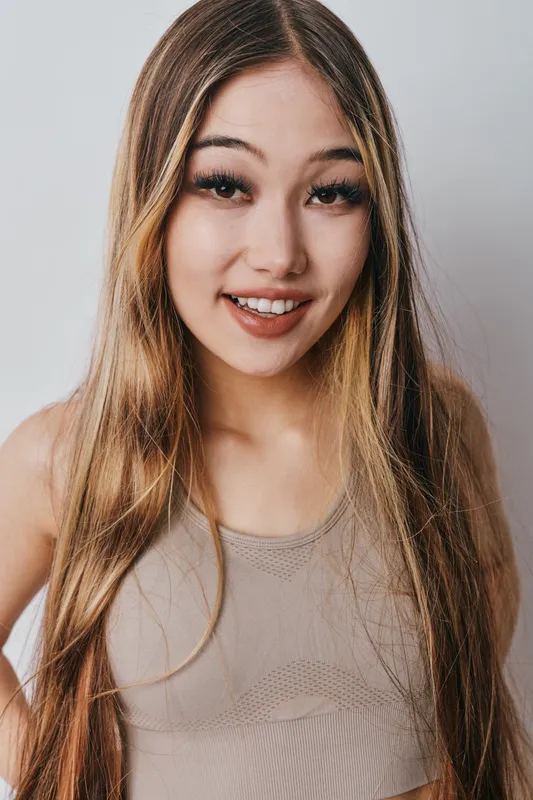 Young woman with long hair smiling wearing beige sleeveless top on plain light background. Portrait of cheerful girl with natural makeup and casual outfit looking at camera.