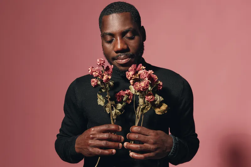 African man holding dried flowers with closed eyes in creative conceptual visual style on pink background. Portrait of thoughtful black male with floral elements in studio.