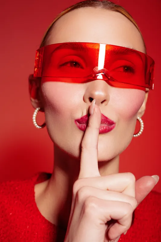 Close-up of a woman wearing red sunglasses and a red sweater making a shushing gesture with her finger on lips. Concept of secrecy, silence, and mystery on red background.
