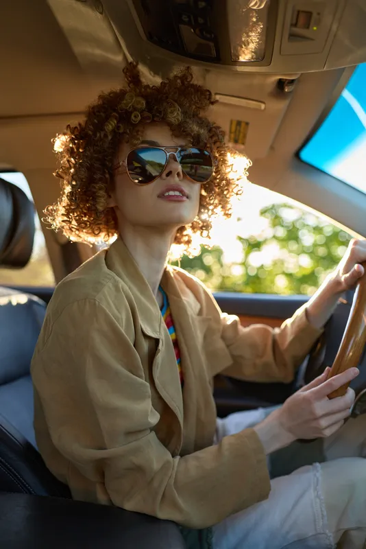 A stylish woman driver in retro tones sits behind the wheel, wearing oversized sunglasses. Natural sunlight filters through the car window, highlighting curly hair and beige attire, vintage mood, and
