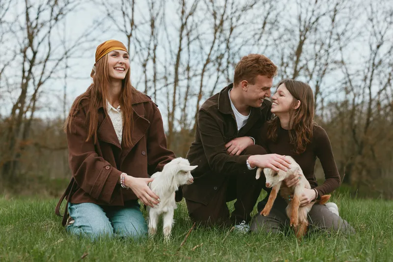 Happy couple outdoors with two small dogs in a grassy park, smiling and interacting, casual jackets and scarves, warm natural light, lifestyle moment celebrating friendship and pets in nature