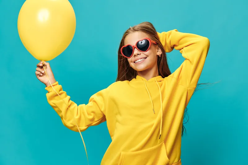 Girl wearing yellow hoodie and red heart sunglasses holding yellow balloon on blue background smiling relaxed casual style youth fun and cheerful mood with long hair