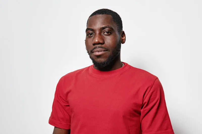 Portrait of confident African American man with dark skin wearing red t shirt standing against white background looking at camera with slight smile and relaxed posture.