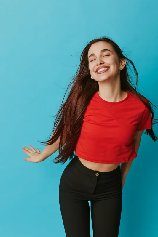Young woman with long hair wearing red crop top and black pants smiling with closed eyes standing against blue background. Casual style, happiness and carefree mood in studio portrait.