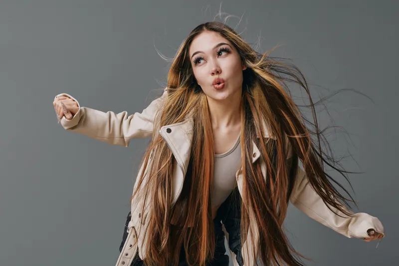 Woman with long hair wearing beige jacket and light shirt posing with expressive face and dynamic posture against grey background in casual style photo shoot.