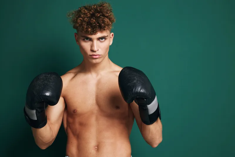 Young male boxer with black gloves posing in fighting stance against green background for fitness, training and sports concept with muscular body.