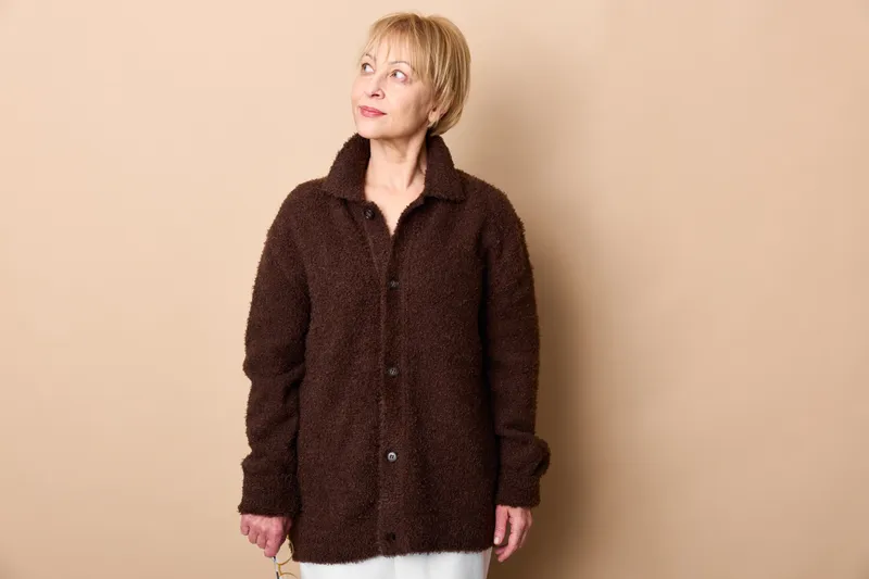 Elderly woman with short hair in dark jacket, standing in studio, beige background. Relaxed portrait with soft light, showing casual fashion and everyday comfort.