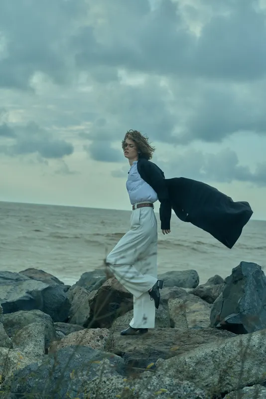Fashion wind coastal sea rocks coat movement silhouette outdoors mood, dramatic windy editorial scene of a woman on rocks with flowing cape and moody sky