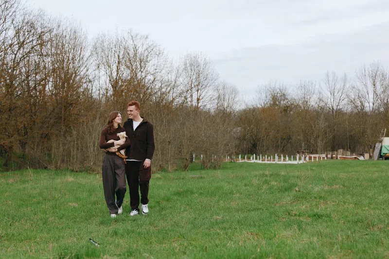 Smiling couple walking in a park, holding a small dog, enjoying a relaxed outdoor moment. Natural daylight and open space convey affection, lifestyle, and companionship with warm daylight.