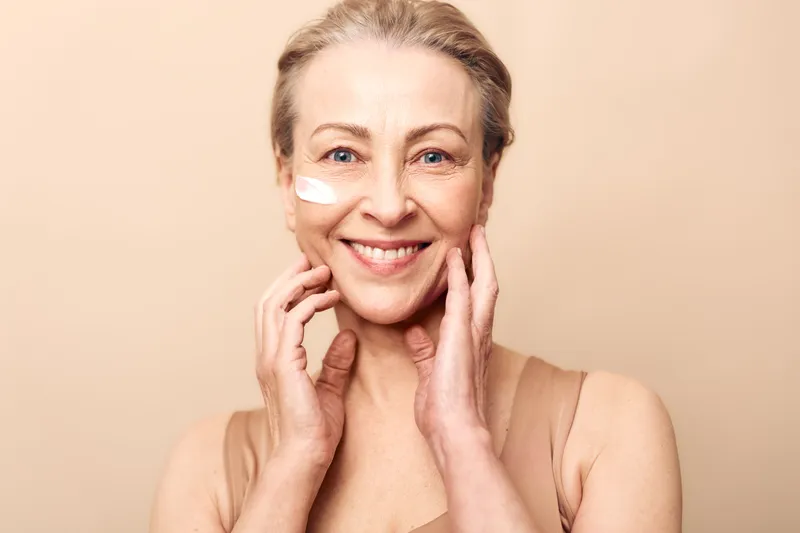 Smiling mature woman with natural skin applies facial cream on cheek, touching face gently. Concept of skincare, antiaging, beauty routine, and selfcare for healthy glowing skin.
