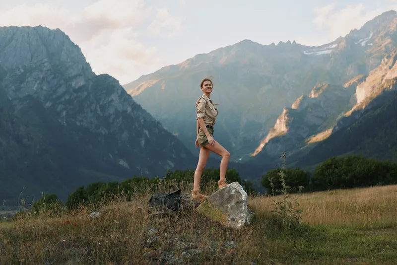 Young woman with short hair stands confidently on a rock in a mountain meadow during golden hour, enjoying the natural landscape and fresh air.