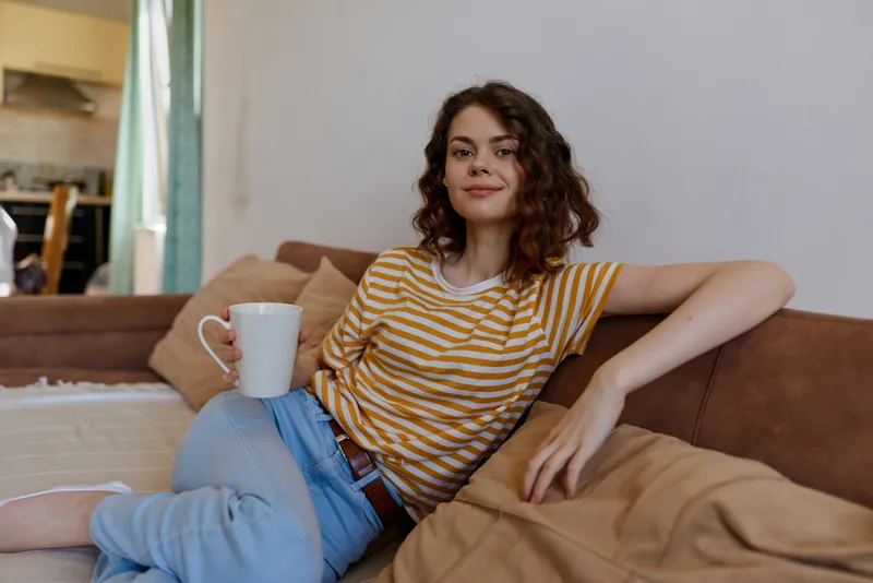 Young woman with curly hair sits relaxed on a cozy couch in a bright living room, holding a mug while wearing a striped shirt for a warm home lifestyle moment.