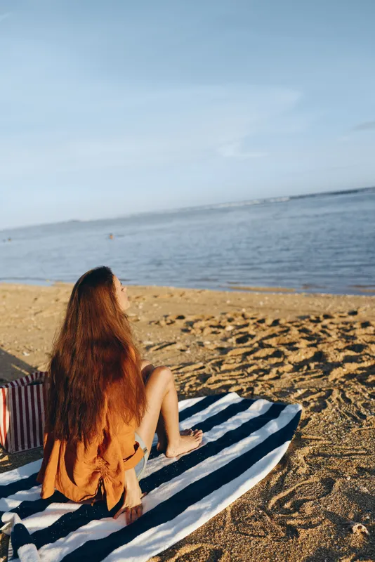 Young woman with long hair sitting on striped towel on sandy beach looking at calm sea under clear blue sky, peaceful moment, warm sunlight, relaxed atmosphere