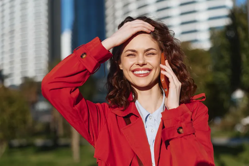 Young woman with curly hair smiles happily while talking on a smartphone outdoors in a city park on a sunny day wearing a red coat and light blue shirt.