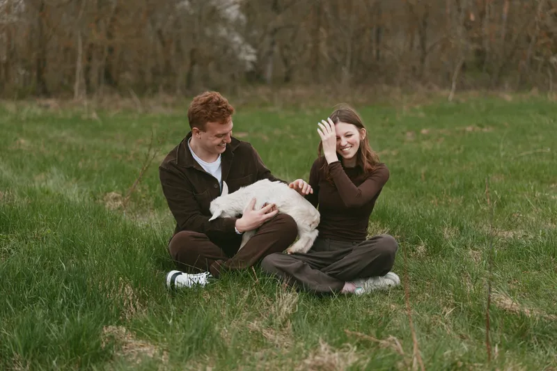 Candid outdoor scene shows a joyful couple in a field with a gentle goat kid, sharing affection and laughter, celebrating nature and togetherness in a rural lifestyle setting.