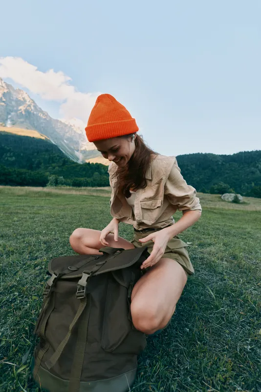 A young woman in an orange beanie and casual clothes kneels on grass unpacking a large backpack in a mountain meadow under clear blue sky. Lifestyle and outdoor adventure concept.
