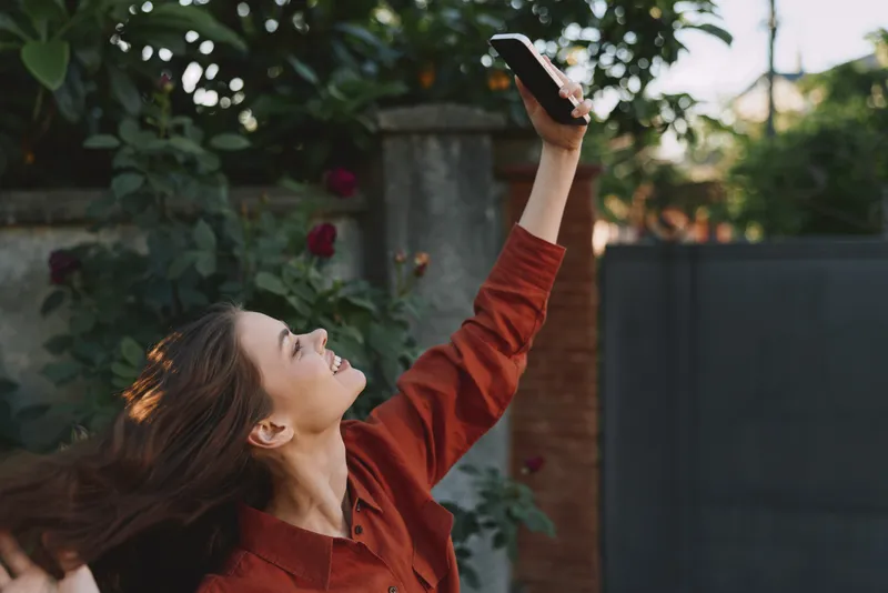 A young woman in a rust sweater raises her phone for a selfie in a sunny garden, smiling as roses and lush greenery frame the moment, conveying casual outdoor joy