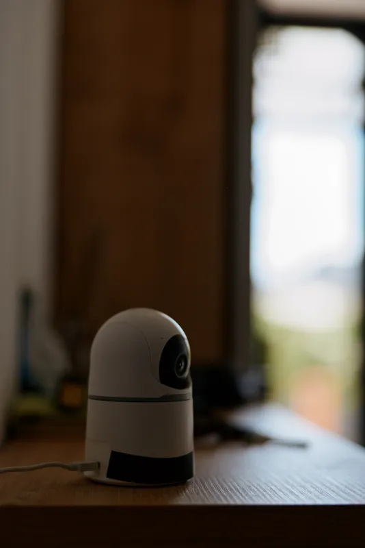 Close up of a small white webcam on a wooden desk in a cozy home interior, soft daylight from a window highlights the device, a modern gadget used for video calls and home security in a minimal