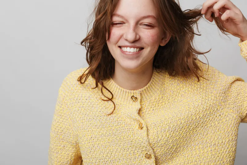 Smiling young woman with curly hair wearing yellow knitted sweater plays with hair on grey background. Natural beauty, casual style, happiness, and carefree mood concept in studio.