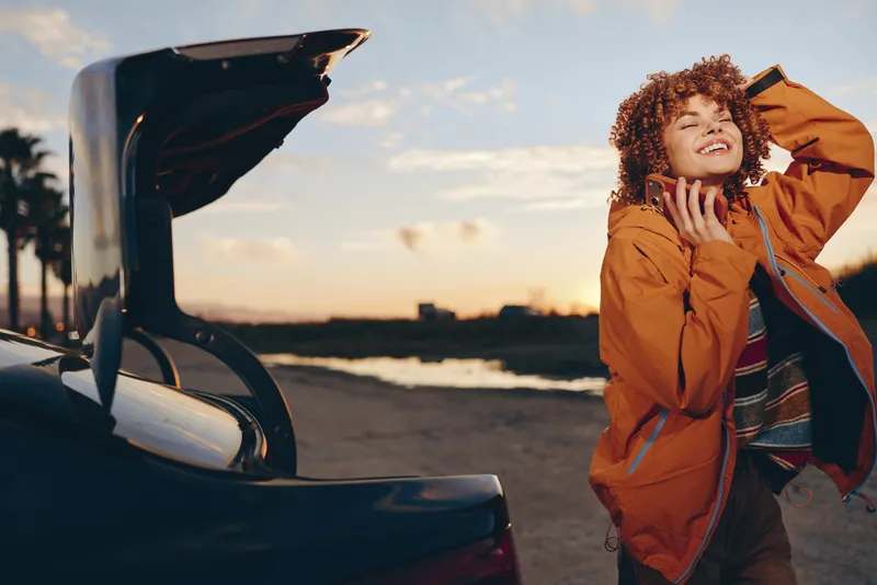 woman, curly, hair, jacket, car, trunk, smile, sunset, lifestyle, outdoor, young woman closes car trunk while chatting and laughing outdoors, warm golden sky, travel mood today