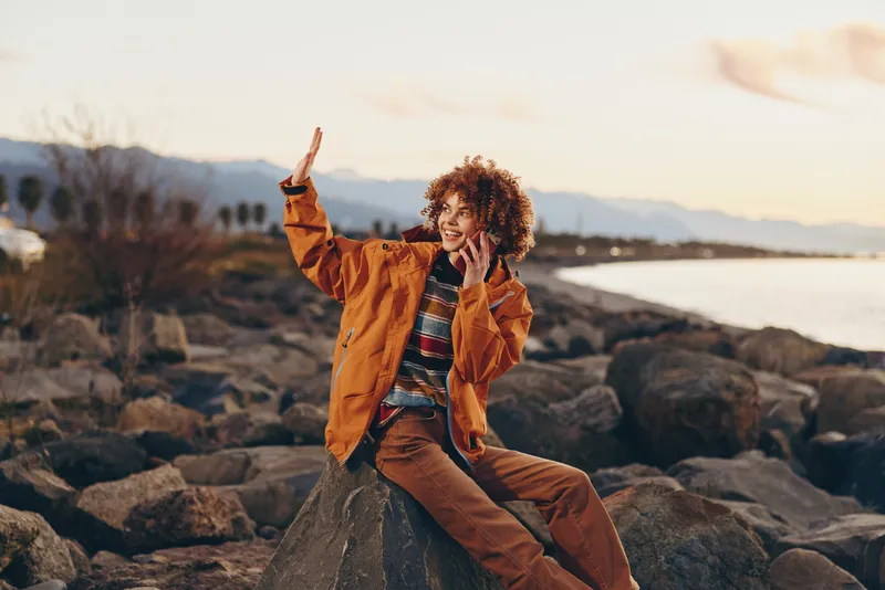Woman lifestyle outdoors by the sea at sunset, standing on rocky shore, warm jacket, curly hair blowing, smiling and laughing, playful gesture, golden light, calm horizon evening