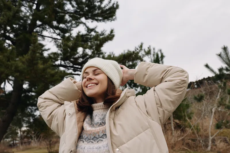 Happy woman in a beige jacket and beanie enjoys a carefree moment outdoors, smiling with closed eyes as trees frame a quiet winter scene.