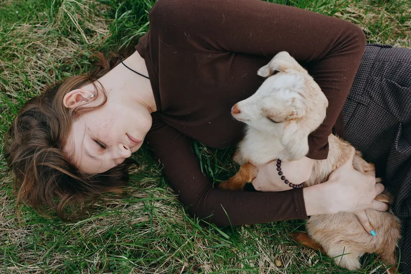 Warm lifestyle scene with a woman lying on grass, embracing a dog, enjoying calm companionship. Natural light, soft textures, wellness and self care in an outdoor moment goat kid.