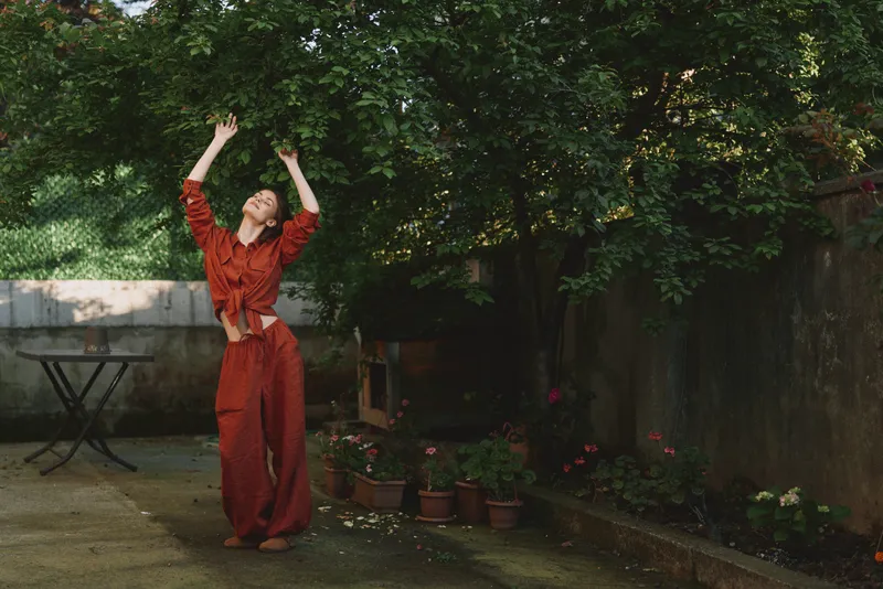 Cheerful woman outdoors, red outfit, dancing pose, sunlit garden scene, joyful mood, relaxed stance, natural colors, lush greenery background